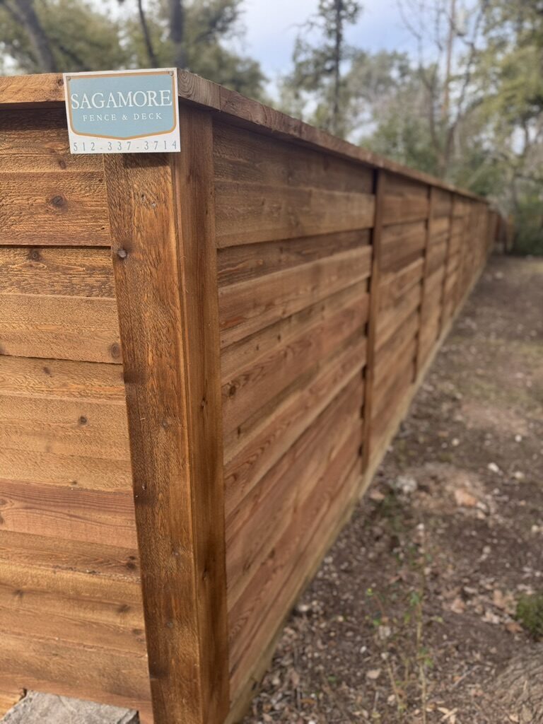 Horizontal cedar wood fence built by Sagamore Fence & Deck, running along a wooded backyard area with a company sign attached.